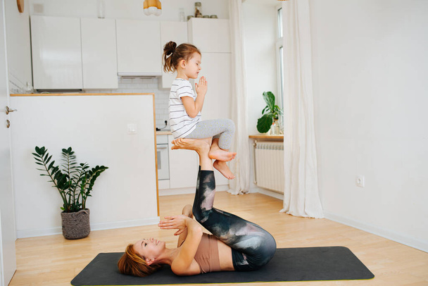 Young mom doing gymnstics with her little daughter on a yoga mat at home. Mom lays on her back, girl sitting on her raised feet like on the chair. Side view. - Foto, imagen