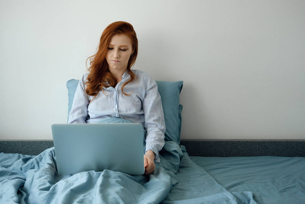 A red-haired girl in her pajamas sits in a blue bed and looks into her laptop with her lips on the side - Photo, image