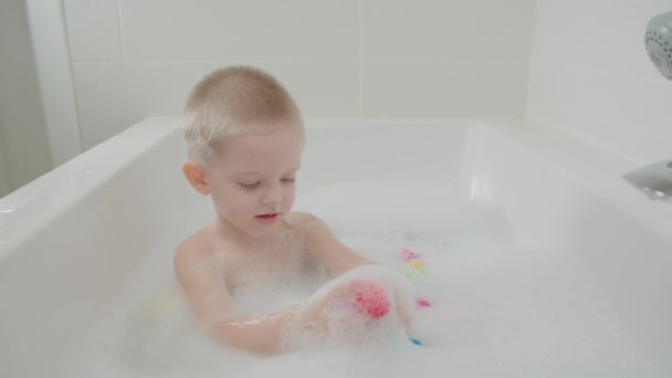 Cheerful boy playing bathroom with foam and toys. Baby with blue eyes and blond - Imágenes, Vídeo