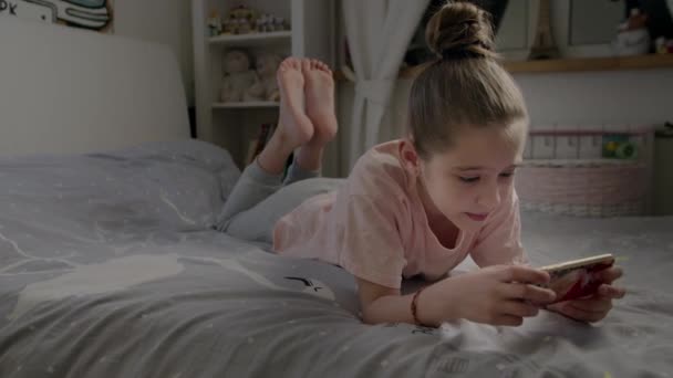 Little girl cute plays in smartphone lying on the bed - Felvétel, videó