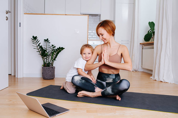 Happy little boy hugs his mother, while doing yoga on a yoga mat in front of a laptop at home. She's looking at her son. She wears tight sportive clothes - Φωτογραφία, εικόνα