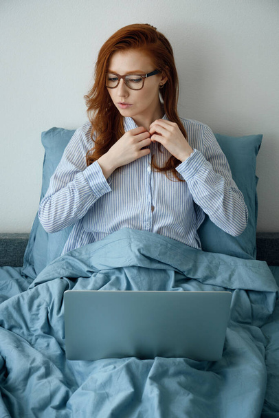 A red-haired girl with glasses sits in bed and adjusts the collar of her shirt, looking at the laptop - Foto, Bild