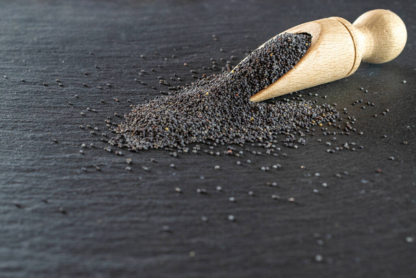 Poppy seeds isolated on dark stone background in wooden scoop. Pile poppyseed food in spoon on black. Healthy breakfast or morning snack diet eating concept. - Fotografie, Obrázek