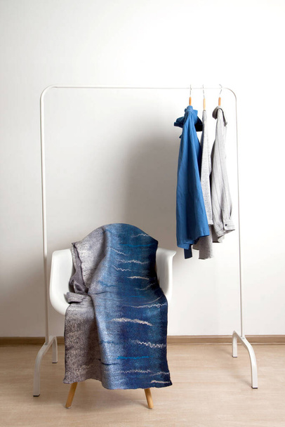 Wonderful gray-blue silk felted designer scarf lies on chair on the background of hanger with shirts on white background in the studio. Stylish homemade clothes concept - Foto, immagini