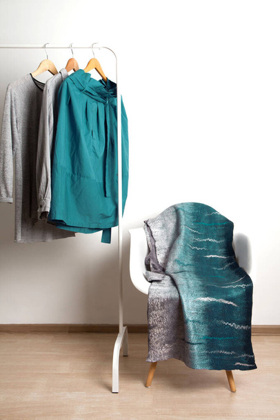 Wonderful gray-blue silk felted designer scarf lies on chair on the background of hanger with shirts on white background in the studio. Stylish homemade clothes concept - Foto, Imagen