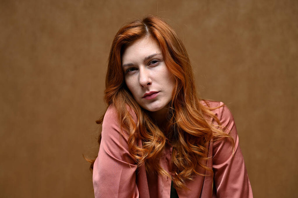 Photo portrait on a beige background of a calm pretty young woman in a pink shirt with long beautiful red hair. - 写真・画像