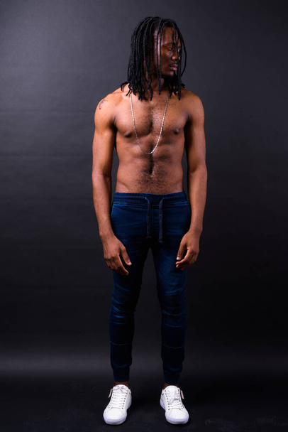 Studio shot of young handsome African man with dreadlocks shirtless against black background - Foto, Imagen