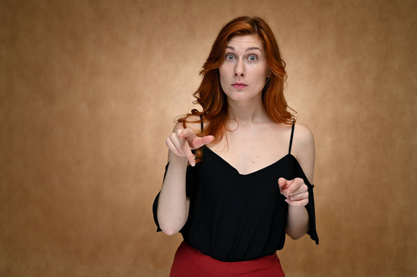 Model actress shows different emotions while talking on camera. Studio photo portrait of a caucasian young woman in a black blouse with long red hair on a beige background. - Valokuva, kuva
