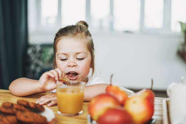 Cute toddler girl with fair hair and big grey eyes eat breakfast at table in bright interior - Foto, afbeelding