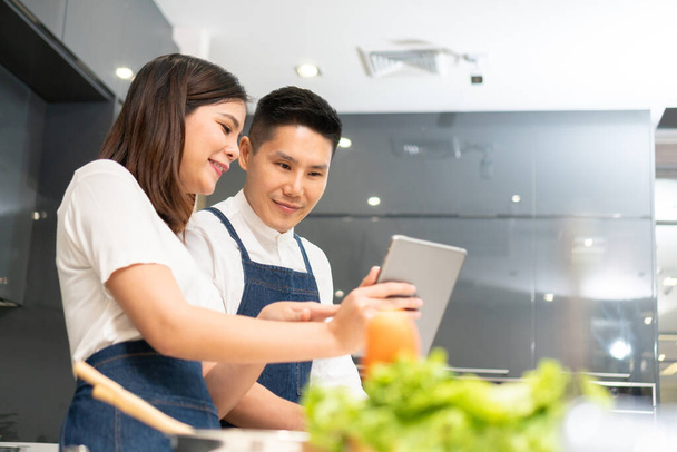 Attractive Smiling Asian couple cooking and using a digital tablet in a modern kitchen at home. Happy Young Man and beautiful woman in white clothes and blue aprons preparing vegetables for the salad - Foto, immagini