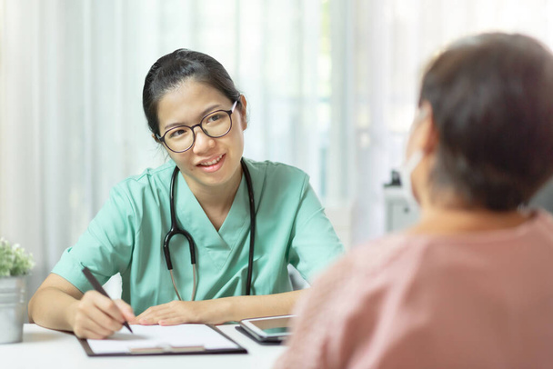 Beautiful Asian Woman Doctor in green uniform wear eyeglasses writing information in paper and talking giving advice to Senior woman patient in medical room at hospital. Disease, Covid 19, Coronavirus - Photo, Image