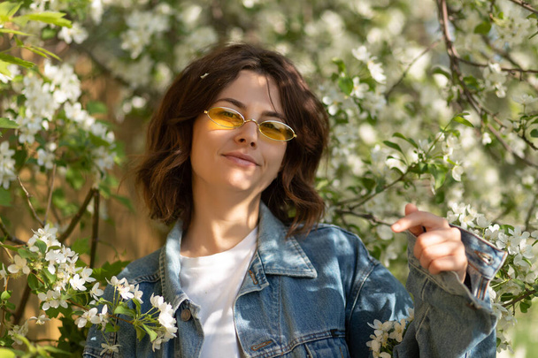 Creative  Portrait woman with flower.Design art concept.romantic portrait of a young woman with curly hair in a jeans jacket with fashionable yellow glasses against the background of a blossoming apple tree derev. 90 style portrait  - Photo, Image