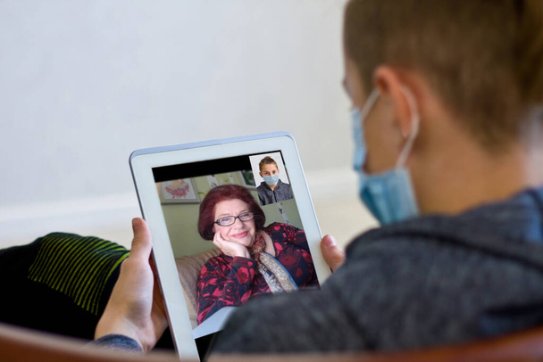 Rear view of a guy talking on a video call with a smiling elderly grandmother, grandson chat online using a mobile device to communicate with a happy grandmother in quarantine at home. - Photo, Image
