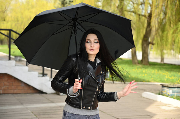 Modelka pózuje proti městskému parku s černým deštníkem v rukou. Jarní venkovní portrét kavkazské módní brunetky dívka v černé bundě a džínách. - Fotografie, Obrázek