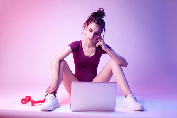 Schlankes Mädchen im Body sitzt auf dem Boden vor weißem Hintergrund in Neonlicht. studiert Online-Training am Laptop - Foto, Bild