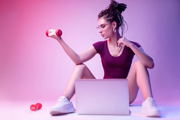 Karcsú lány testőruhában ül a padlón, fehér háttérben neon fényben. tanulmányok online képzés egy laptop - Fotó, kép