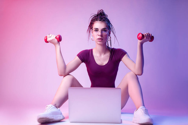 slender girl dressed in a bodysuit sits on the floor on a white background in neon light. studies online training on a laptop - Foto, Imagem