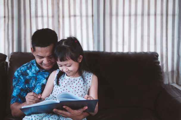 Μικρό κορίτσι και ο πατέρας απολαμβάνουν ανάγνωση του βιβλίου μαζί στο σπίτι.Κοινωνική απόσταση κατά τη διάρκεια της καραντίνας, Online έννοια της εκπαίδευσης - Φωτογραφία, εικόνα
