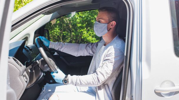 Quarantine concept. Pandemic. A young guy-taxi driver in a gray protective medical mask and a gray jacket is sitting behind the driver's seat in the car. He looks straight. Virus. Coronavirus. Insulation  - Foto, imagen