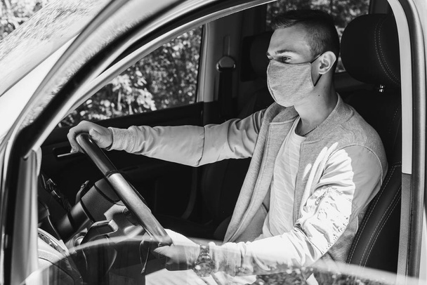 Karantén koncepció. Pandémia. Egy fiatal taxisofőr szürke orvosi maszkban és szürke dzsekiben ül a vezetőülés mögött, egyenesen előre néz. Koronavírus. Fekete-fehér fénykép - Fotó, kép
