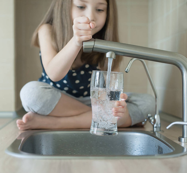 Little girl open a water tap with her hand holding a transparent glass. Kitchen faucet. Glass of clean water. Filling cup beverage. Pouring fresh drink. Hydration. Healthcare. Healthy lifestyle - Foto, afbeelding