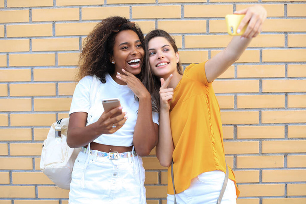 Two young smiling hipster women in summer clothes posing on street.Female showing positive face emotions - Foto, afbeelding