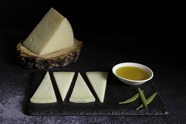 čtyři plátky typického španělského sýra manchego a mísy olivový olej na břidlicovém talíři a kus sýra manchego na tmavém pozadí. Kopírovat prostor - Fotografie, Obrázek