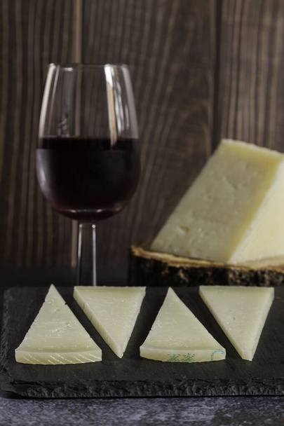 平板の上の典型的なスペインのマンチェゴチーズ4切れ、木製の背景の木のスライス上の赤ワイングラスとピースマンチェゴチーズ。スペースのコピー - 写真・画像