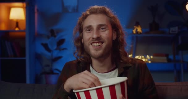 Close up view of young cheerful man watching funny show while eating popcorn. Handsome guy with earrings and long hair laughing while sitting on couch in front of tv. Concept of leisure. - Footage, Video