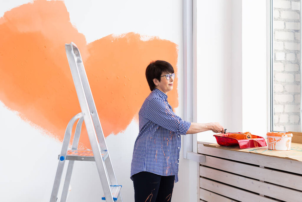 Szczęśliwa kobieta w średnim wieku maluje ściany wewnętrzne wałkiem malarskim w nowym domu. Kobieta z wałkiem malującym ścianę. - Zdjęcie, obraz