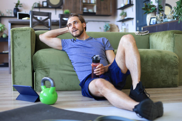 Sporty man resting, having break drinking water after doing exercise sitting on floor in living room. Fintess at home during self isolation - Foto, immagini