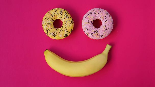 Two donuts and a banana lie on pink surface, forming a smile emotion - Φωτογραφία, εικόνα
