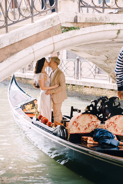 Casamento Itália em Veneza. Um gondoleiro rola uma noiva e um noivo em uma gôndola de madeira clássica ao longo de um estreito canal veneziano. Gondola flutua sob uma ponte de pedra, os recém-casados se beijam
. - Foto, Imagem