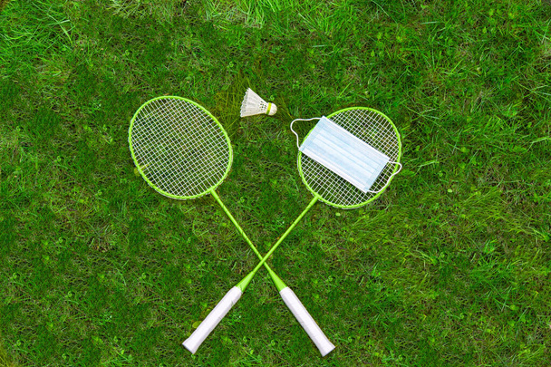 Coronavirus protection sing. Badminton rackets and shuttlecock on green grass. Medical mask on one racket. view from above. ban - Photo, Image