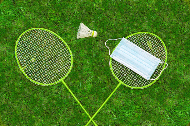 Coronavirus protection symbol. Badminton rackets and shuttlecock on green grass. Medical mask on one racket. view from above. forbiddance - Fotografie, Obrázek
