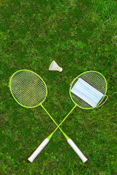 Coronavirus protection symbol. Badminton rackets and shuttlecock on green grass. Medical mask on one racket. view from above. embargo - Foto, Imagem