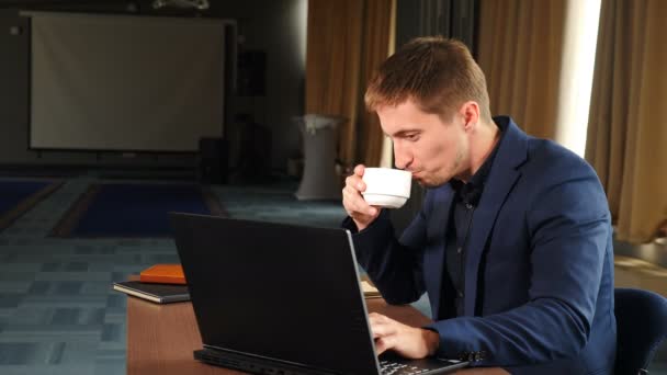 Fiatal üzletember az irodában laptoppal dolgozik az irodában, mosolyog kávézás gépelés a számítógépen szörfözés web nézi képernyőn élvezi a munkát. 4 k felvétel - Felvétel, videó
