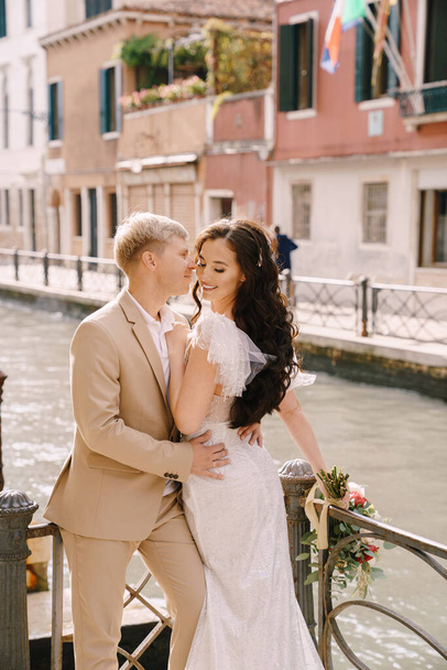 ヴェネツィアでのイタリア結婚式。新婚旅行は、ヴェネツィア運河の銀行を採用立っている。新郎は花嫁を腰で抱きしめる。小さな美しい列車と砂色のメンズスーツと白いウェディングドレス. - 写真・画像