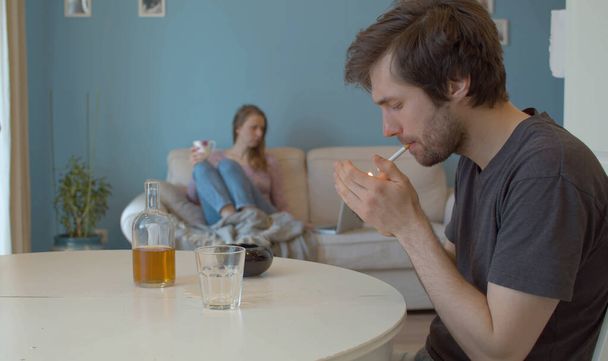 Ένας μεθυσμένος, λυπημένος άνθρωπος ανάβει τσιγάρο. Στο βάθος, στο defocus, η σύζυγος κάθεται στον καναπέ και κοιτάζει τον υπολογιστή. - Φωτογραφία, εικόνα