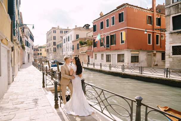 Italy wedding in Venice. Newlyweds stand embracing on the banks of the Venice Canal. The groom hugs the bride by the waist. White wedding dress with small beautiful train and sand-colored mens suit. - Photo, Image