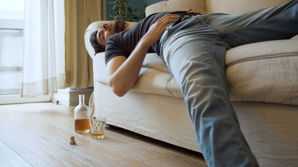 Opilý muž spí na gauči oblečený. Nedaleko je nedopitá láhev whisky a sklenička. Mladý debauchee ve svém bytě po večírku - Fotografie, Obrázek