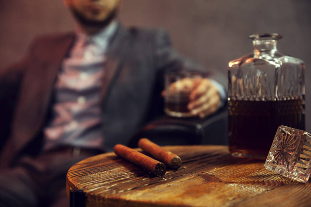Two cigars and a carafe of whisky on wooden table and an elegant man with the glass of whisky in the background - Foto, imagen