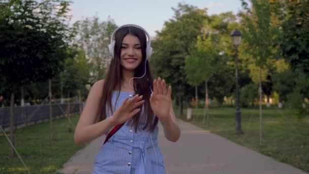 Dancing brunette woman in white headphones. Attractive lady is using technologies in her everyday life, having fun. Spending time in the park, outdoors in summer.  - Video