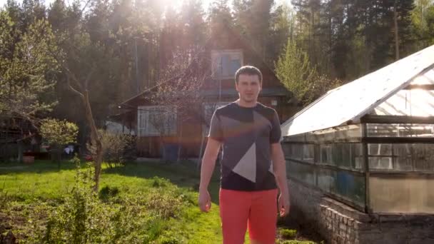 Fröhlicher erwachsener Mann im Garten gegen Gewächshaus und Landhaus im Dorf - Filmmaterial, Video