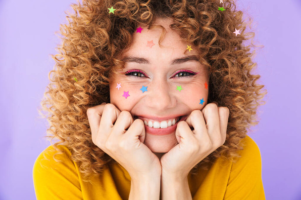 Image closeup of attractive curly girl having fun and smiling with multicolored glitter stars on her face isolated over violet background - Foto, Imagem