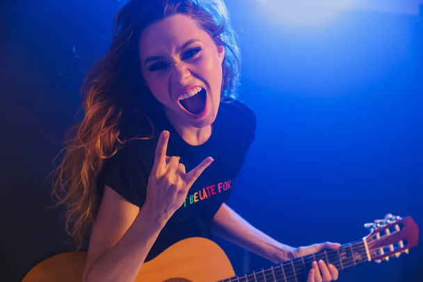 Image of emotional amazing young positive woman musician play on a guitar on scene in night club over dark background with flash lights showing rock gesture. - Фото, изображение