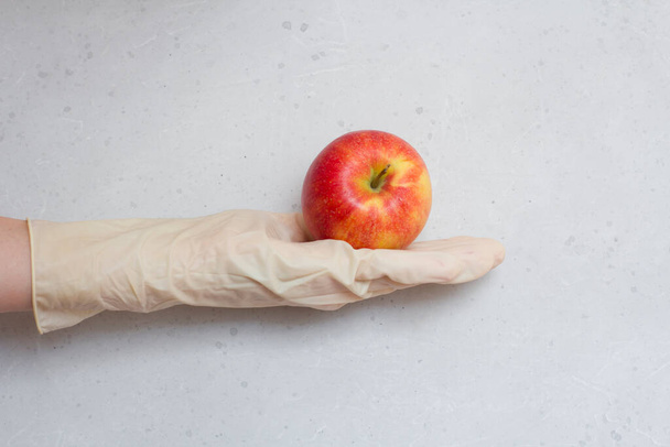 Manzana roja se encuentra en la mano en guante de látex de goma desechable blanco, sobre un fondo de hormigón moderno gris claro y blanco. Una mano en un guante de goma sostiene una fruta de manzana. Concepto epidémico de Coronavirus
. - Foto, Imagen