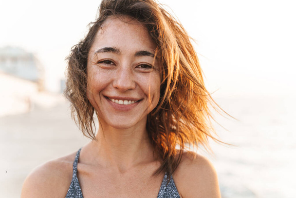 Portrait Nahaufnahme einer fröhlichen Frau in Sommerkleidung, die lächelt und in die Kamera schaut, während sie morgens am Meer spaziert - Foto, Bild