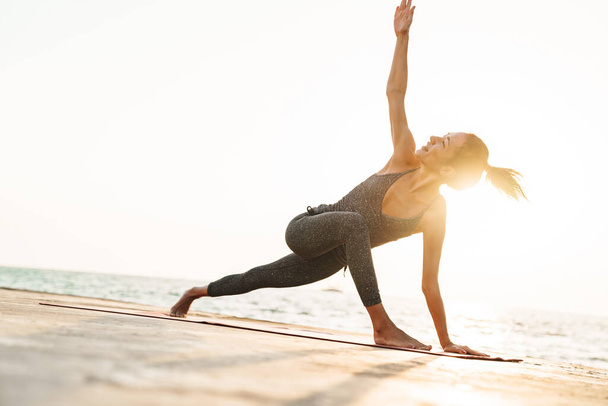 Photo of happy sportswoman in tracksuit stretching her body on mat while practicing yoga by seaside in morning - Valokuva, kuva