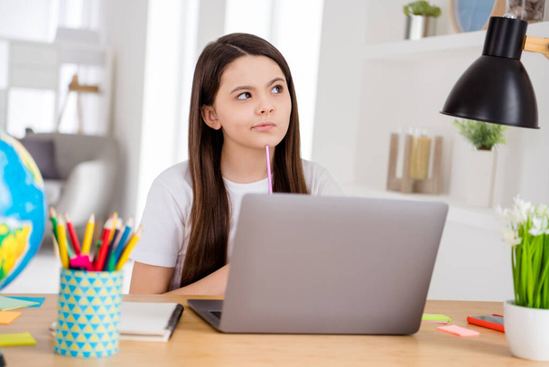 Photo of little school girl notebook online lesson video conference listen teacher minded question pencil on chin sit table desk distance quarantine study comfort room classroom indoors - Foto, Imagen